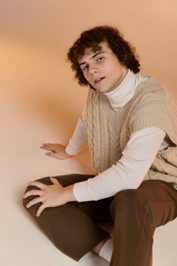 Kıvırcık saçlı genç bir çocuk samimi bir stüdyoda örülmüş bir yelek giyiyor.