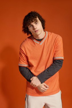 Genç kıvırcık saçlı bir çocuk, turuncu renklerle çevrili, rahat bir şekilde poz verir..