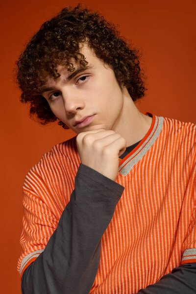 A young boy with curly hair stands thoughtfully, dressed in casual attire, showcasing unique style.