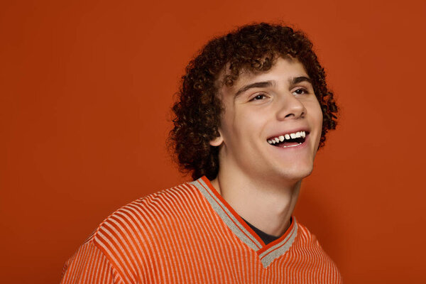 A young boy with curly hair smiles brightly while wearing casual clothing in orange.
