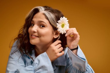 Beyaz perçemli ve şık kıyafetli genç bir kadın stüdyoda poz verirken gülümsüyor..