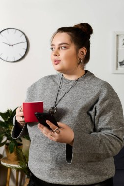 Güzel, büyük beden bir kadın telefonunu kontrol ederken elinde bir bardak tutuyor, kendine güven ve sıcaklık saçıyor..