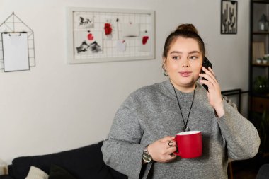 Süslü bir süveter giymiş iri yarı bir kadın telefonda konuşurken elinde kırmızı bir bardak tutuyor..