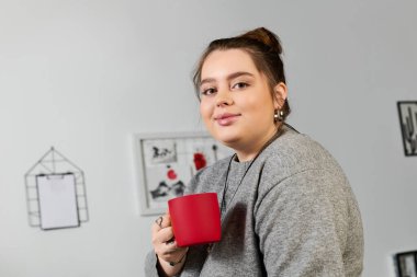 Güzel ve iri bir kadın şık bir odada kırmızı bardaktan yudumlarken rahatlıyor..