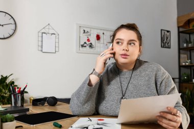 Artı boyutta bir kadın, yaratıcı bir işte çalışıyor, ofisinde telefonla konuşurken elinde kağıt tutuyor..