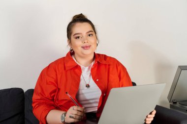 Güzel ve iri yarı bir kadın rahat bir ev ortamında dizüstü bilgisayarıyla çalışırken gülümsüyor..