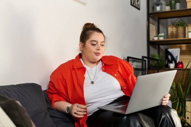 Güzel ve iri yarı bir kadın rahat bir şekilde kanepeye oturur, rahat bir yaşam alanında dizüstü bilgisayarına odaklanır..