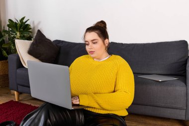 Güzel ve iri yarı bir kadın, rahat bir oturma odasında yerde otururken dizüstü bilgisayarına odaklanmış..