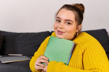 Rahat bir atmosferde, güzel ve iri bir kadın gülümser, göğsüne bir defter tutarak..