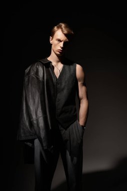 Yakışıklı genç bir adam, loş bir alanda kendine güvenerek duruyor, modern erkekliğini sergiliyor..