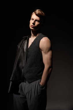 Genç bir adam yeleğine cazibe ve güven saçar. Elinde deri bir ceket tutar..