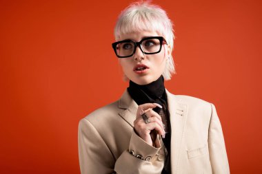 Gözlüklü ve büyük beden ceketli kendinden emin genç bir kadın canlı turuncu bir arkaplana karşı poz veriyor..