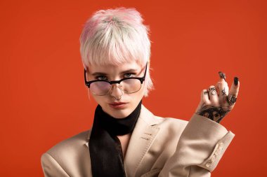 Kısa pembe ve beyaz saçlı, gözlüklü ve dövmeli kendine güvenen genç bir kadın cazibe yayar..