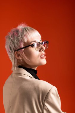 Gözlüklü ve modern saç kesimli kendine güvenen genç bir kadın parlak turuncu bir arka plana gülümsüyor..