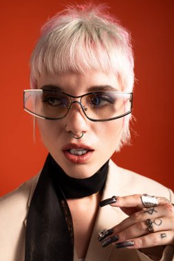 Gümüş saçlı, şık gözlükleri olan genç, zarif bir kadın ateşli kırmızı bir fonda duruyor..