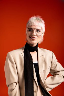 Şık giyinmiş, şık gözlükler takmış kendine güvenen genç bir kadın cesur turuncu bir arka planda duruyor..