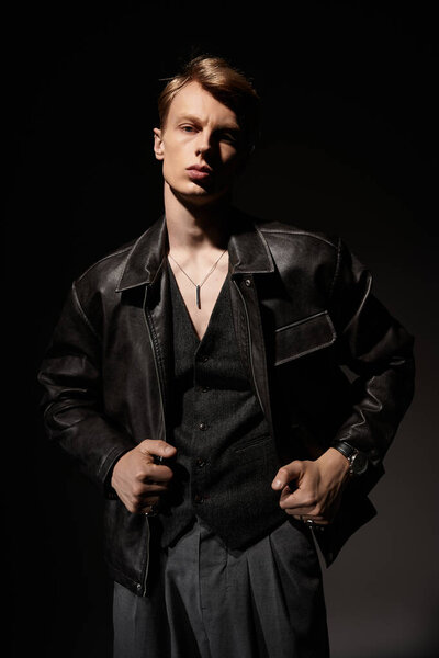A young and handsome man stands confidently in a dark setting wearing a chic leather jacket.