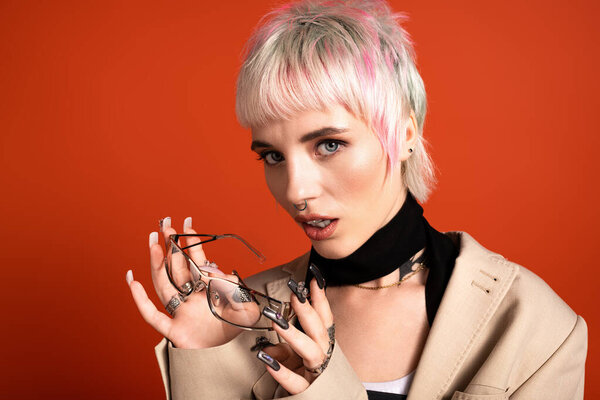 Молодая женщина с яркими волосами уверенно позирует в стильных очках на оранжевом фоне.