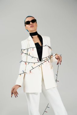 Şık beyaz takım elbiseli genç bir kadın, renkli kış ışıklarıyla çevrili, kendinden emin poz veriyor..