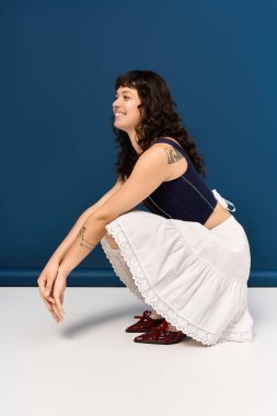 Çekici genç kadın, benzersiz tarzını sergileyen modaya uygun bir kıyafetle çömelirken gülümsüyor..