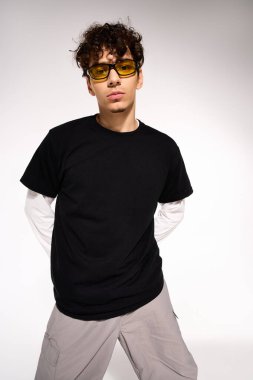 A young man with curly hair and sunglasses poses confidently in a studio, showing off a modern look.