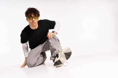 Kıvırcık saçlı ve güneş gözlüklü genç bir adam şık bir stüdyo ortamında yaratıcı bir poz veriyor..