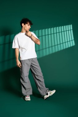 Kıvırcık saçlı bir genç, modaya uygun giyinip, stüdyoda modayı ön plana çıkarıyor..