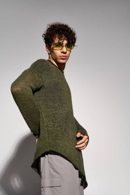 Şık bukleli genç bir adam stüdyoda poz verirken güneş gözlüğü takıyor..