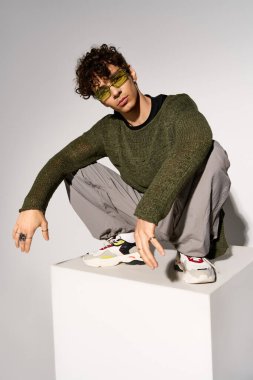 A young man with curly hair poses stylishly in sunglasses, showing off his outfit in a studio.