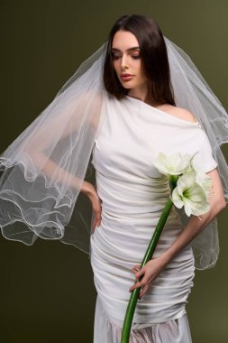 Genç gelin zarif bir şekilde elinde büyük bir çiçek, eşsiz gelinlik güzelliğini ve tarzını sergiliyor..