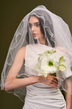 Genç bir gelin zarif bir elbiseyle duruyor, peçesi yüzünü çerçeveliyor ve elinde çiçekler.