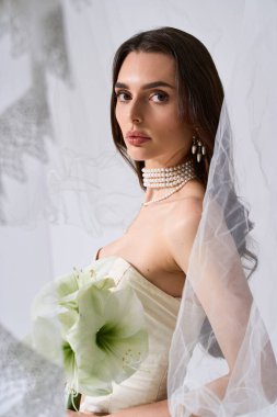 Genç gelin elinde buketle zarifçe poz veriyor, yumuşak ışıkta zarafeti somutlaştırıyor..