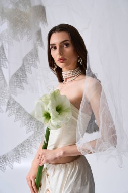 Parlak gelin zarif bir şekilde ayakta duruyor, elinde bir buket çiçekle karışık peçesini gösteriyor..