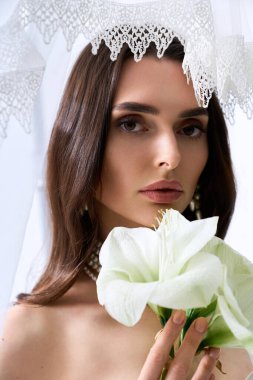 Göz kamaştırıcı genç bir gelin zarif bir şekilde elinde beyaz bir çiçek, saf neşe saçarak ayakta duruyor..