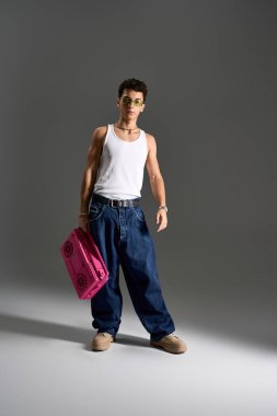 Beyaz kolsuz tişörtlü ve güneş gözlüklü şık bir genç, kasetçalar ile gri bir arkaplana yaslanıyor..