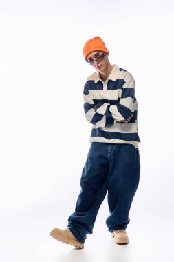 Eğitimli bir genç, çizgili polo ve turuncu bir şapkayla beyaz bir arka plana karşı güvenle poz verir..