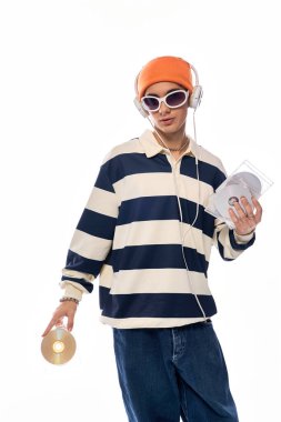 Bir genç, çizgili uzun kollu polo ve turuncu şapkayla kendinden emin bir şekilde poz verir..