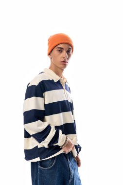 Genç, çizgili uzun kollu polo ve turuncu şapkayla beyaz zemine karşı güvenle duruyor..