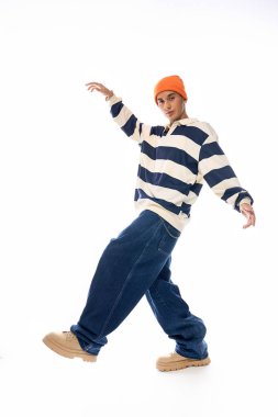Çizgili uzun kollu polo ve turuncu şapkalı bir genç, beyaz arka planda güvenle poz veriyor..