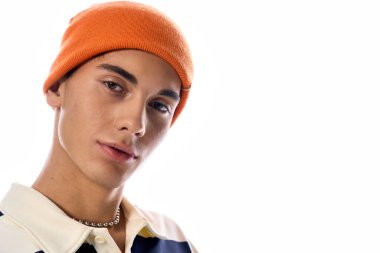 Genç, çizgili uzun kollu polo ve turuncu şapkayla beyaz bir arkaplanda güven içinde poz veriyor..