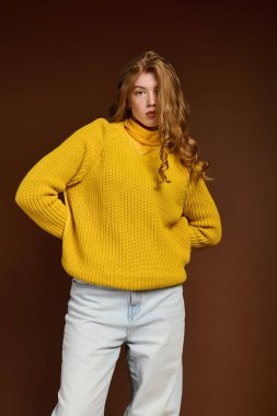 Kızıl saçlı genç bir kadın rahat sarı bir süveterle kahverengi bir fona karşı güvenle poz veriyor..