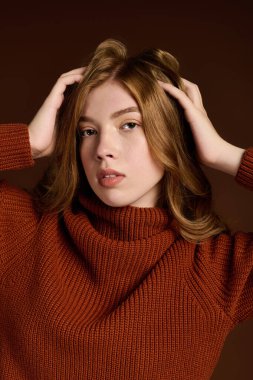 Göz kamaştırıcı kızıl saçlı genç bir kadın kahverengi bir arka fona karşı şık bir kış süveteri içinde poz veriyor..