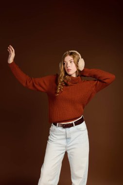 Kızıl saçlı genç bir kadın, kahverengi bir arka plana kış süveteri giyip zarif bir poz veriyor..