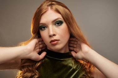 Göz kamaştırıcı bir kızıl saçlı, gri bir stüdyoda poz veriyor, cesur göz makyajı yapıyor ve şık bir şekilde..