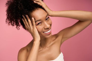 Genç kadın neşeyle gülümsüyor, yumuşak pembe renklerle kaplanmış, mutluluk saçıyor..