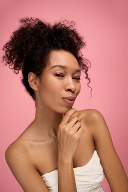 Kendinden emin genç bir kadın, çenesi elinde, pembe bir arka planda usulca gülümsüyor..