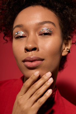 Genç kadın zarif bir şekilde poz veriyor, göz kamaştırıcı sanatsal makyajını kırmızı bir zemine vurguluyor..