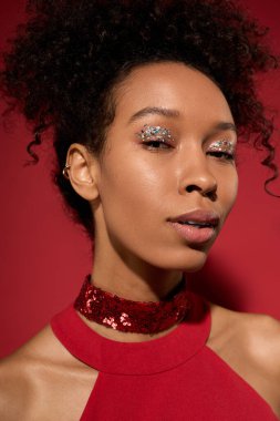 Genç bir kadın zarif bir şekilde gülümserken göz kamaştırıcı makyajını ve şık kıyafetlerini kırmızıyla gösterir..