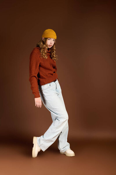 A confident young redhead woman poses in a cozy sweater and beanie against a brown backdrop.