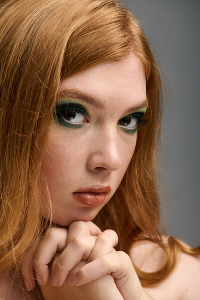 Красивая рыжая женщина изящно позирует, демонстрируя поразительный макияж глаз в серой студии.